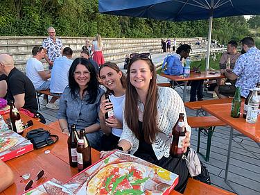 Drei Frauen sitzen an einem Biertisch am Eckert Beach, lachen in die Kamera und halten Bierflaschen in der Hand; vor ihnen liegen offene Pizzakartons, im Hintergrund weitere Gäste in geselliger Runde.