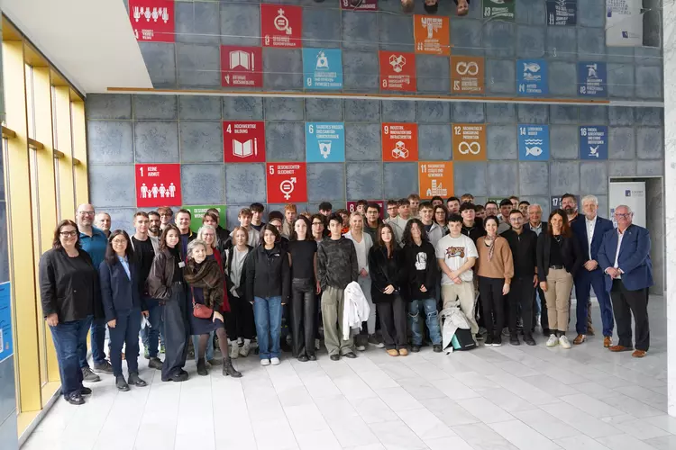 Teilnehmende sitzen in einem hellen Raum und hören einem Vortrag zu, der von vorne gehalten wird; eine spiegelnde Decke reflektiert die Szene.