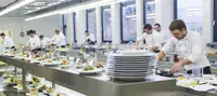 Prüfungsvorbereitung in der Profiküche – angehende Küchenmeister im Praxiseinsatz Auszubildende in weißen Kochjacken arbeiten in einer großen Profiküche an verschiedenen Gerichten, die auf einer langen Edelstahlfläche zur Bewertung angerichtet sind.