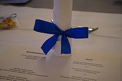 Zusammengerollte Stoffserviette mit blauer Schleife, daneben eine Menükarte mit Speise- und Weinfolge.