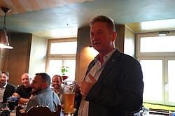 Ein Mann hält bei einem gemeinsamen Abendessen eine Ansprache, während mehrere Teilnehmende aufmerksam zuhören.