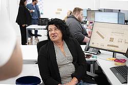 Eine Frau sitzt an einem Büroschreibtisch vor einem Bildschirm. Sie unterhält sich mit anderen Teilnehmern.