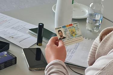 Eine Person hält einen Ausweis in der Hand. Auf dem Tisch liegen Unterlagen, ein Smartphone, eine UV-Lampe und weitere Arbeitsmaterialien. Das Bild zeigt eine praktische Übung zur Prüfung von Dokumenten im Rahmen einer Schulung zur Dokumentensicherheit. Bild 2: