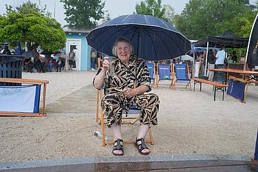 Eine ältere Frau sitzt bei Regen auf einem Strandstuhl, hält einen Regenschirm über sich und lächelt in die Kamera. In der Hand hat sie ein Glas mit Getränk, im Hintergrund sind weitere Sitzplätze und Gäste zu sehen.
