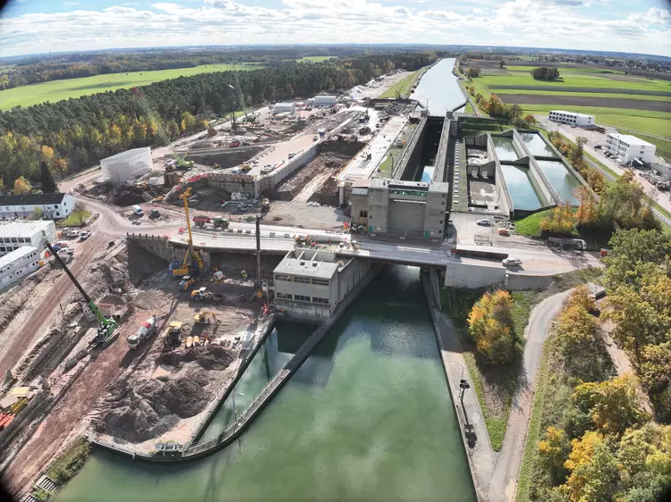 Luftaufnahme einer weitläufigen Baustelle an einer Schleusenanlage mit Wasserstraße, Baumaschinen und angrenzenden Feldern und Waldflächen.
