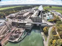 Luftaufnahme einer weitläufigen Baustelle an einer Schleusenanlage mit Wasserstraße, Baumaschinen und angrenzenden Feldern und Waldflächen.