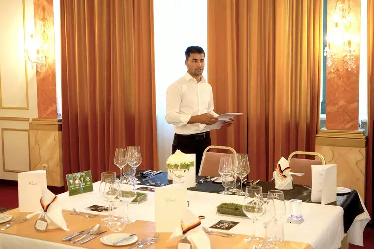 Gastlichkeit mit Konzept Ein junger Mann im weißen Hemd steht in einem festlich eingedeckten Restaurant, hält Unterlagen in der Hand und spricht, vermutlich bei einer Präsentation im Rahmen der Hotelfachschulausbildung.