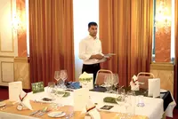 Gastlichkeit mit Konzept Ein junger Mann im weißen Hemd steht in einem festlich eingedeckten Restaurant, hält Unterlagen in der Hand und spricht, vermutlich bei einer Präsentation im Rahmen der Hotelfachschulausbildung.
