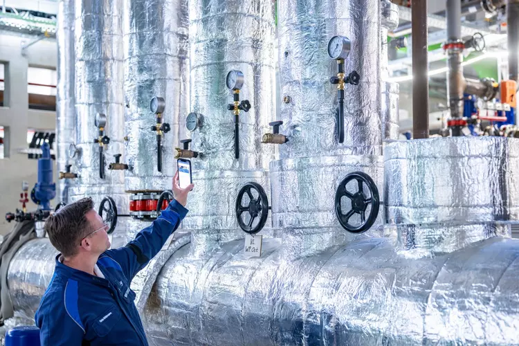 Technische Präzision in der Praxis Ein Fachmann überprüft die Anzeigen und Ventile einer verrohrten, silbrig glänzenden Industrieanlage mithilfe eines digitalen Geräts. Das Bild steht für die Verbindung aus technischem Know-how, Digitalisierung und modernster Messtechnik.