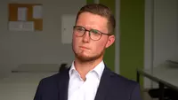 Junge Mann mit dunkelbonden kurzen Haaren und Brille blickt relativ ernst in die Kamera. Er trägt Bart, eine blaue Jacke und ein weißes Hemd. 