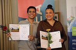 Zwei Absolventen stehen nebeneinander, halten ihre Urkunden und Blumen und lächeln in die Kamera.