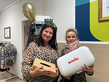 Fairschenkladen Brotbox Zwei Kolleginnen stehen im Fairschenkladen. Beide mittlerern Alters, eine schwarzhaarig die andere blond. Die linke Frau hält eine hölzerne Brotbox ind er Hand, die rechte Dame hält eine weiße Brotbox mit dem Nutella Logo in der Hand. Beide stehen nebeneinander und freuen sich über ihren Fund.