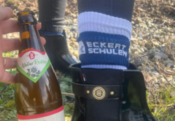 Eine Person hält eine Flasche Bischofshof Natur Radler in der Hand. Im Vordergrund sind schwarze Gummistiefel zu sehen, aus denen weiße Socken mit dem Aufdruck Eckert Schulen hervorblitzen. Der Hintergrund zeigt Wiese und Kiesboden – ein Hinweis auf die Pflanzaktion im Freien.