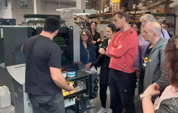 Gruppe beobachtet Demonstration einer Offsetdruckmaschine durch einen Mitarbeiter in einer Druckerei.