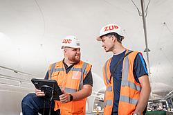 Zwei Baufachkräfte mit Schutzhelm und Warnweste besprechen auf einer Baustelle Inhalte auf einem Tablet.