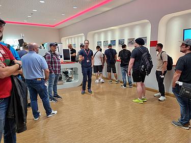 Gruppe von IT-Exkursionsteilnehmenden im Showroom mit technischen Geräten und Unternehmenspräsentation.