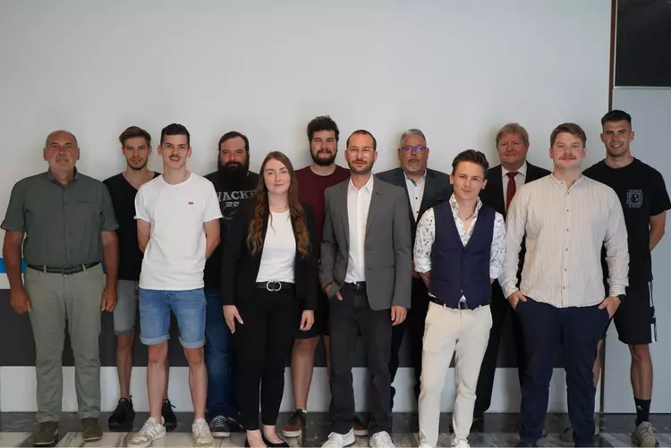 Gruppenfoto bei der Projektbesprechung Gruppenfoto von sechs Personen in unterschiedlicher Business-Kleidung vor einer weiß gestrichenen Wand mit dekorativen Farbstreifen. Alle stehen nebeneinander und lächeln in die Kamera.