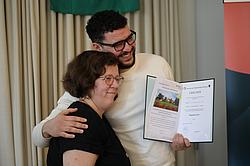 Zwei Personen stehen eng beieinander, halten eine Urkunde und lächeln in die Kamera während sie sich umarmen.
