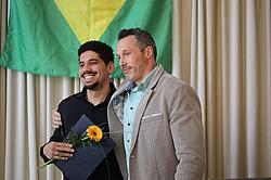 Zwei Männer stehen vor einer brasilianischen Flagge, einer legt dem anderen den Arm um die Schulter. Der Geehrte hält eine Urkunde und eine Blume in der Hand und lächelt.