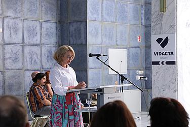 Ein Grußwort mit Herz Eine Frau mit blonder Kurzhaarfrisur steht an einem Rednerpult mit Mikrofon, trägt eine weiße Bluse und einen bunten Rock. Im Hintergrund sitzen Zuhörende. Rechts ist ein Schild mit der Aufschrift „VIDACTA Schulen“.