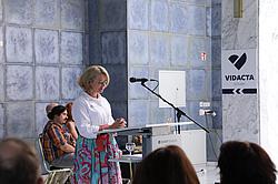 Eine Frau mit blonder Kurzhaarfrisur steht an einem Rednerpult mit Mikrofon, trägt eine weiße Bluse und einen bunten Rock. Im Hintergrund sitzen Zuhörende. Rechts ist ein Schild mit der Aufschrift „VIDACTA Schulen“.