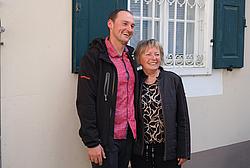 Ein Absolvent posiert gemeinsam mit einer älteren Frau vor einem Gebäude und lächelt in die Kamera.