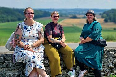 Drei Teilnehmerinnen sitzen auf einer Steinmauer vor einer hügeligen Landschaft mit Feldern und Wäldern.