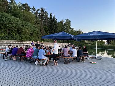 Zwei lange Biertische mit ehemaligen Schüler:innen der MG-Klasse auf einem Holzdeck am Eckert Beach. Die Gruppe sitzt unter großen dunkelblauen Sonnenschirmen, umgeben von Bäumen und Wasser bei untergehender Sonne.