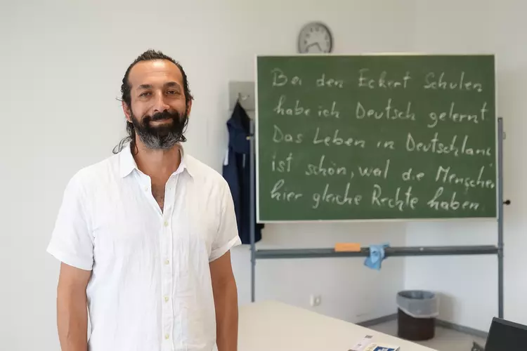 Teilnehmer eines Deutschkurses an den Eckert Schulen steht lächelnd im Unterrichtsraum vor einer Tafel mit deutschem Text.