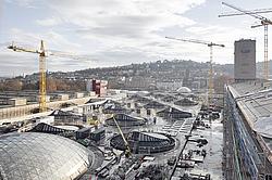 Blick über eine Großbaustelle mit mehreren Baukränen, Dachkonstruktionen und einem Rohbau in einer Stadt.