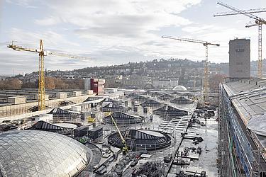 Großbaustelle in einem städtischen Umfeld mit mehreren Baukränen, Hallendächern und Gebäuden im Hintergrund.