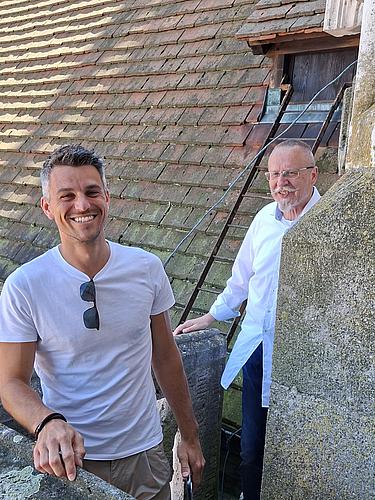 Zwei Männer stehen zwischen steinernen Mauerteilen auf dem Dachbereich eines historischen Gebäudes. Einer lächelt direkt in die Kamera, der andere blickt zur Seite. Im Hintergrund ist ein altes Dach mit verwitterten roten Ziegeln und eine kleine Dachluke mit Metallleiter zu sehen.