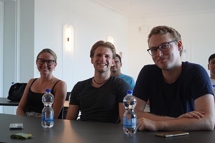 Mit Motivation und Teamgeist ins neue Semester Drei junge Menschen – zwei Männer und eine Frau – sitzen nebeneinander in einem Seminarraum, lächeln in die Kamera. Auf dem Tisch stehen Wasserflaschen. Im Hintergrund sind weitere Teilnehmende zu sehen.