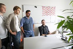 Vier Männer befinden sich in einem Büro. Während ein Mann spricht hören die anderen drei aufmerksam zu.