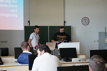 Techniker der Eckert Schulen präsentieren ihre Projektarbeit im Klassenzimmer. Zwei Männer stehen vor der Leinwand und halten einen Vortrag. Weitere Studierende sitzen im Klassenzimmer und hören gespannt zu. 