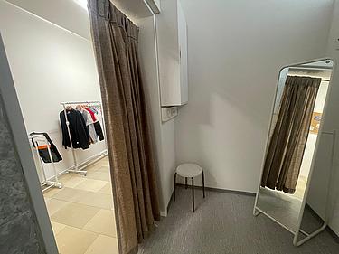 Die Umkleidekabine im Fairschenkladen. Der Vorhang ist beige und leicht geöffnet. In der Umkleidekabine steht ein kleiner Hocker in der Ecke und ein Spiegel.