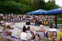Livemusik mit Seeblick – Sommerkonzert am Eckert Beach Eine Band spielt auf einer Bühne am Wasser. Zu sehen sind Musiker mit Keyboard, Percussion und Gitarre unter blauen Sonnenschirmen. Viele Menschen sitzen auf Liegestühlen oder den Steinstufen des Amphitheaters, das dicht mit Publikum gefüllt ist. Im Hintergrund dichter Wald.