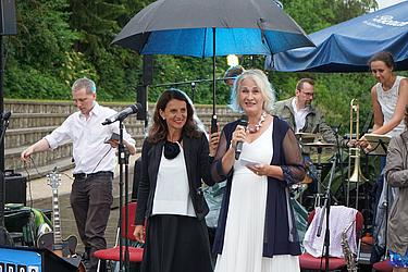 Zwei Frauen stehen bei Regen auf einer Bühne und sprechen ins Mikrofon. Eine von ihnen hält einen großen Regenschirm. Im Hintergrund sieht man Musikinstrumente und Musiker.