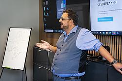 Ein Mann referiert vor einem kleinen Whiteboard und einem Bildschirm im Hintergrund. Der Mann artikuliert sich mit seinen Händen.