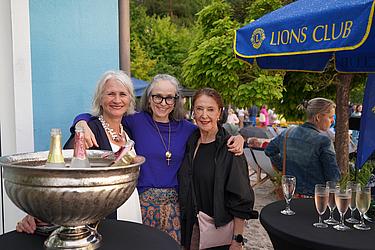 Drei Frauen stehen lachend nebeneinander an einem Stehtisch mit einer großen Schale voller Sektflaschen. Im Hintergrund sieht man weitere Gäste, einen Baum und einen Lions Club-Schirm.