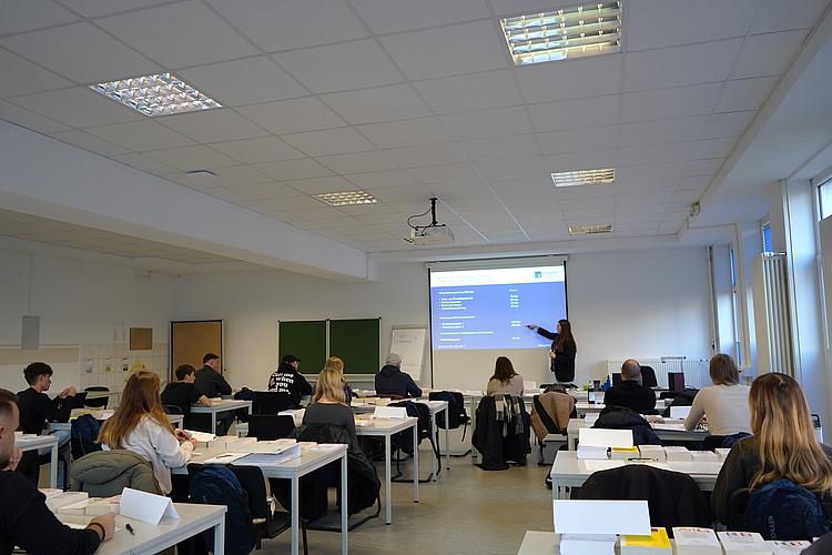 Wirtschaftsfachwirt Kursstart Regensburg Eckert Schulen Ein heller Unterrichtsraum an den Eckert Schulen in Regensburg: Mehrere Teilnehmerinnen und Teilnehmer sitzen an Tischen und folgen einer Dozentin, die vorne am Beamer eine Präsentation zum Wirtschaftsfachwirt zeigt.