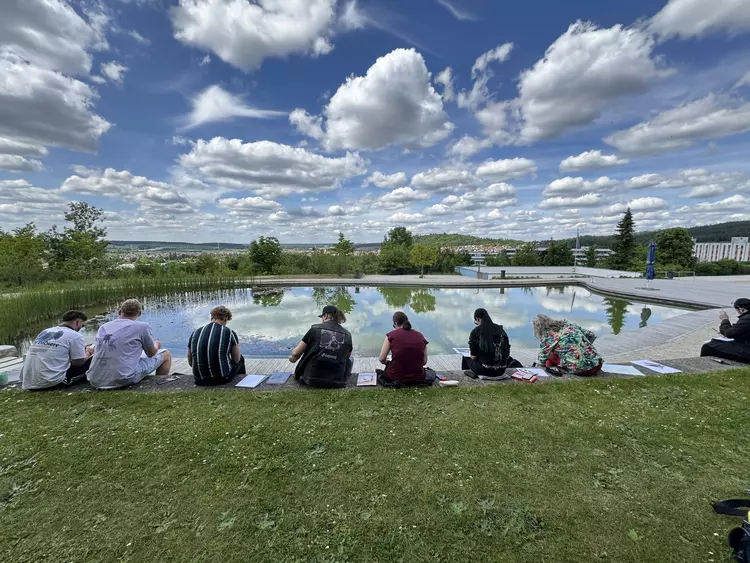 Studierende der Kreativ-Gruppe sitzen am Teich des Campus Regenstauf und arbeiten bei sonnigem Wetter an Zeichnungen und Projekten.