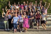 25 Jahre MG02 Großes Gruppenbild mit rund 40 fröhlichen Personen, die mit erhobenen Armen auf einer Treppe im Freien posieren – sonniger Tag, lockere Kleidung, ausgelassene Stimmung.
