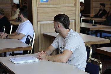Ein Student sitzt in einem großen Saal der Eckert Schulen an einem Tisch, schaut nach links und hört aufmerksam zu. Vor ihm liegt ein aufgeschlagenes Notizbuch.