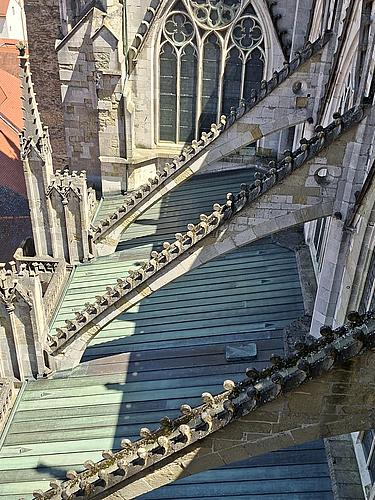 Schräg von oben fotografierte gotische Strebebögen am Regensburger Dom. Die steinernen Bögen mit dekorativen Aufsätzen stützen die hohen Außenmauern. Darunter ein grünlich oxidiertes Kupferdach. Im Hintergrund sind spitzbogige Fenster mit Maßwerk zu sehen.