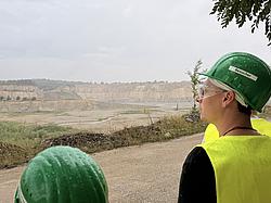 Blick über den weitläufigen Kalksteinbruch mit hellen, terrassenförmigen Abbauwänden; im Vordergrund zwei Studierende mit grünen Besucherhelmen und Schutzbrille.