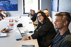 Die Gruppe hört einer Frau zu, die ebenfalls mit um den Tisch sitzt. Die Frau hat ihren Laptop offen.