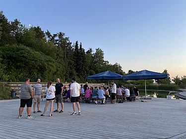 Abendliche Szene am Eckert Beach mit mehreren Gruppen ehemaliger Schüler:innen, die auf dem Holzdeck stehen oder an Tischen unter großen Sonnenschirmen zusammensitzen. Im Hintergrund spiegelt sich die untergehende Sonne im Wasser.