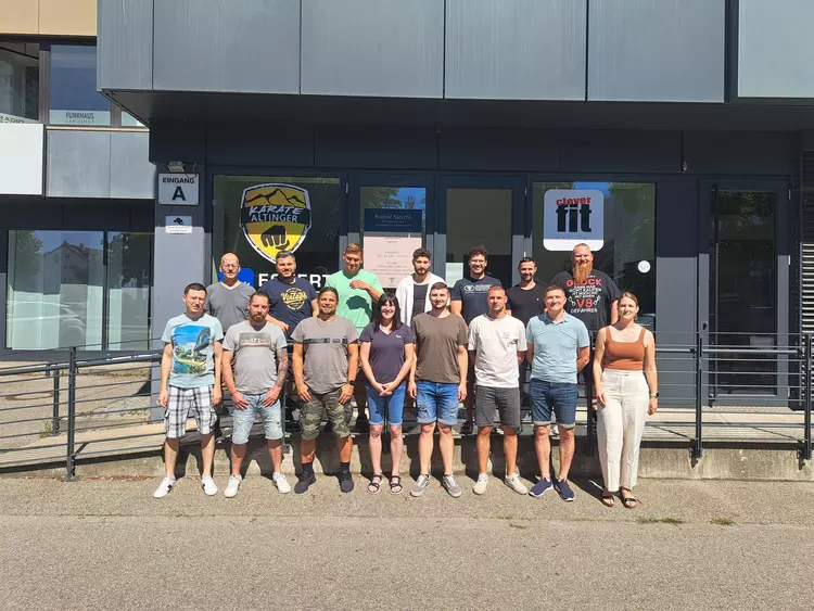 Abschluss mit Teamgeist – Gruppenfoto vor dem Trainingszentrum Eine Gruppe von 18 Personen – Männer und Frauen – steht in zwei Reihen für ein Gruppenfoto vor einem modernen Gebäude mit Glasfassade. Hinter ihnen sind die Logos „Karate Altinger“ und „Clever Fit“ sichtbar. Die Stimmung ist freundlich und sommerlich.