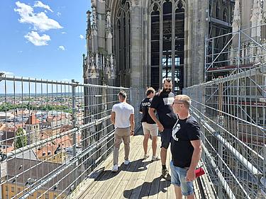 Mehrere Männer stehen auf einem eingerüsteten Steg in großer Höhe am Regensburger Dom. Umgeben von Schutzgittern blicken sie auf die Stadt und die architektonischen Details der gotischen Kathedrale. Im Hintergrund ist ein weiter Blick über die Dächer von Regensburg zu sehen.